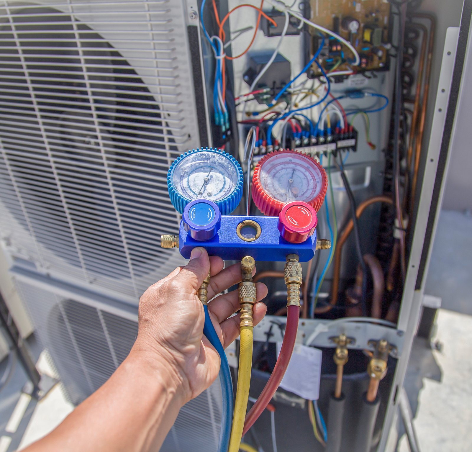 HVAC technician using a manifold gauge to check refrigerant levels on an outdoor air conditioning unit with exposed wiring and components.